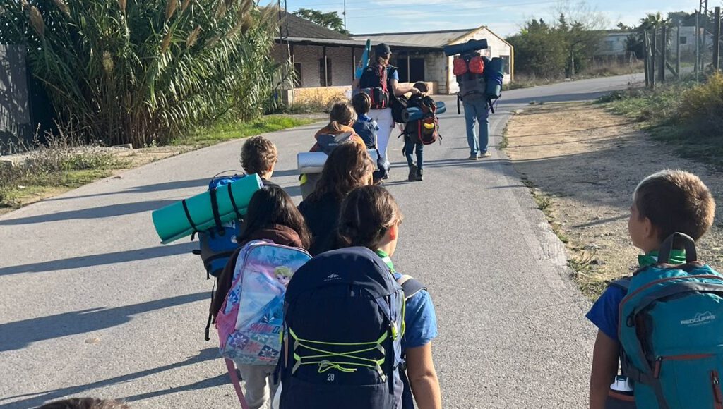 Grupo de niños scouts caminando por una carretera con mochilas y esterillas, dirigiéndose a un campamento.