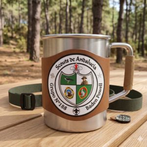 Taza metálica de camping con asa y funda de cuero, decorada con el logo de Scouts de Andalucía del Grupo Scout 188 Baden Powell, colocada sobre una mesa en un entorno de bosque.
