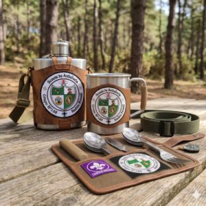 Set de campamento scout compuesto por cantimplora metálica con funda de cuero, taza de camping y cubiertos en funda enrollable, todos con el escudo de Scouts de Andalucía del Grupo Scout 188 Baden Powell, sobre una mesa en un entorno de bosque.