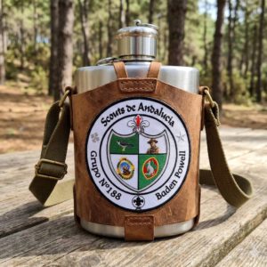 Cantimplora metálica con funda de cuero y correa, decorada con el escudo de Scouts de Andalucía del Grupo Scout 188 Baden Powell, colocada sobre una mesa en un entorno de bosque.
