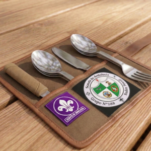 Juego de cubiertos de camping guardado en una funda enrollable, acompañado de parches de Scouts de Andalucía y del Grupo Scout 188 Baden Powell, sobre una mesa de madera en un entorno natural.