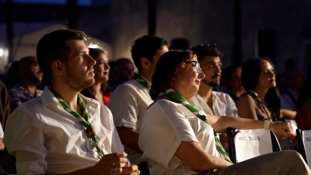 Grupo de scouts adultos sentados entre el público durante el acto de celebración del 55 aniversario, llevando el pañuelo verde y blanco del grupo mientras observan el documental.