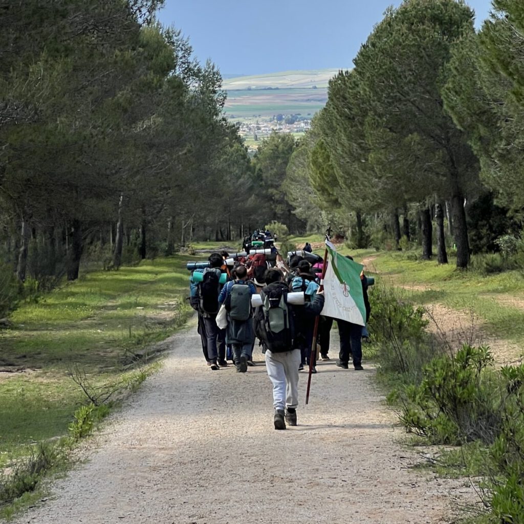 Grupo de scouts caminando con mochilas y la bandera del grupo por un camino forestal rodeado de árboles durante una ruta en plena naturaleza.